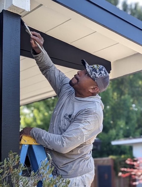 A person wearing a gray long-sleeve shirt and a camouflage cap is standing on a blue stepladder, painting the underside of an outdoor structure with a brush. The person is focused on the task and is surrounded by greenery.