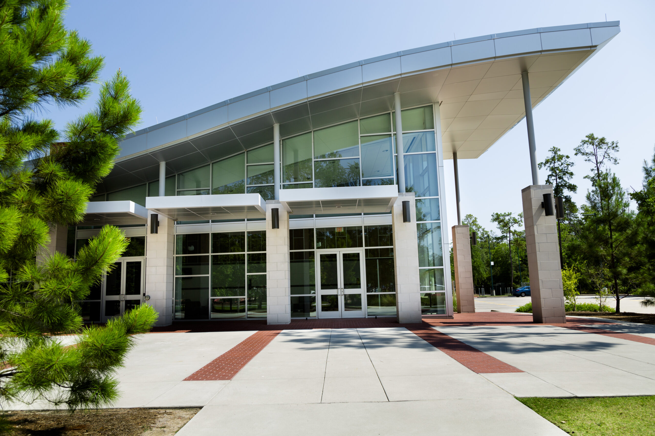 A modern building with a large glass facade and an overhanging roof stands surrounded by trees. The entrance features wide glass doors, and the foreground shows a paved walkway bordered by red bricks. The sky is clear and blue.