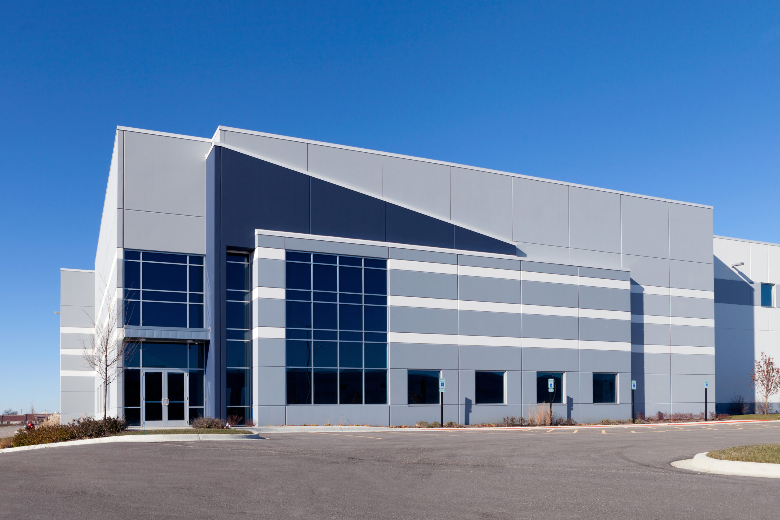 A large, modern industrial building with a grey facade and rows of dark-tinted windows. The structure is set against a clear blue sky, with an empty parking lot and a few small trees in the foreground.