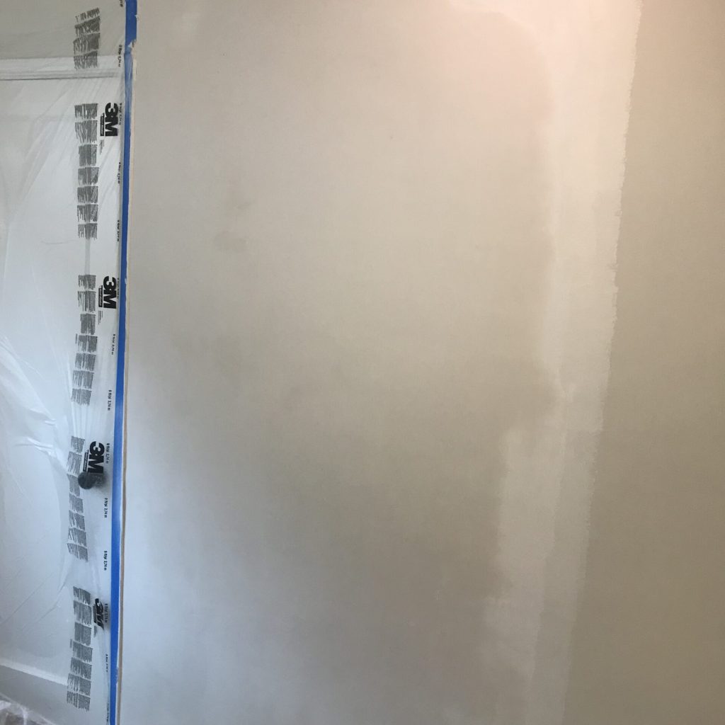 A partially painted wall with sections taped off for precision, displaying progress in a room renovation. electrical outlets and painting supplies are also visible.