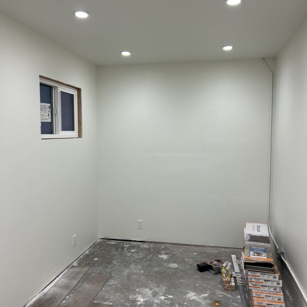 A small room under construction with gray flooring partially installed. tools and flooring materials are scattered on the floor. the room features white walls, recessed lighting, and one visible window.
