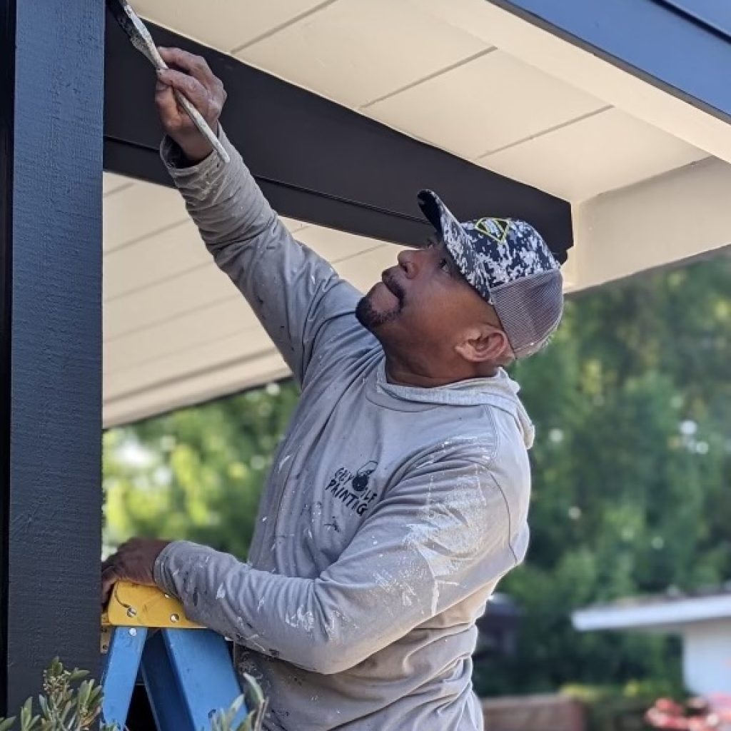 A person wearing a gray long-sleeve shirt and a camouflage cap is standing on a blue stepladder, painting the underside of an outdoor structure with a brush. The person is focused on the task and is surrounded by greenery.
