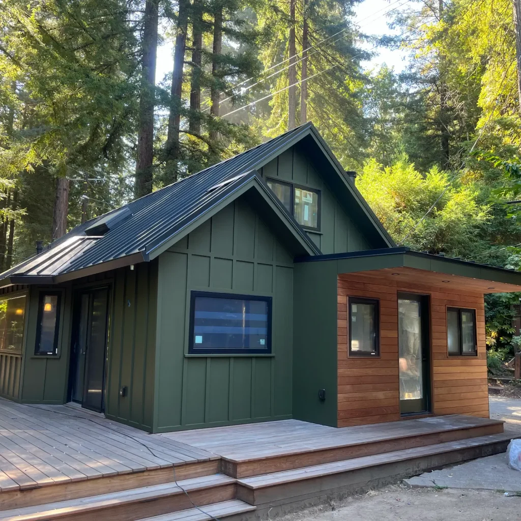 Green painted cabin with wooden deck in forest setting.