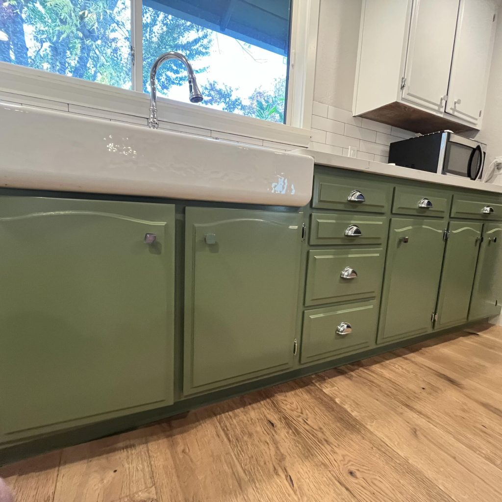 A kitchen featuring green cabinets with silver handles, a white farmhouse sink, and a hardwood floor treated with interior paint, viewed from a low angle.